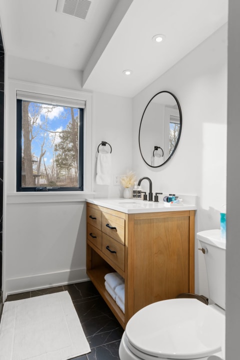 Modern bathroom with wooden vanity, round mirror, and large window.