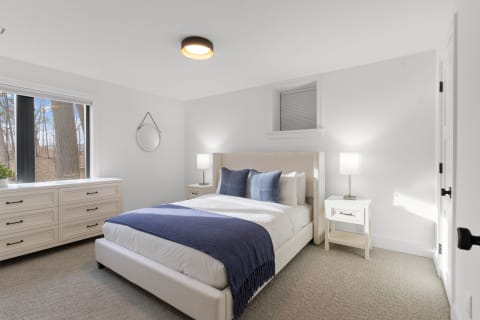 A modern bedroom with a white bed, blue throw blanket, and a window view of trees.