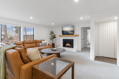 Modern living room with leather sofa, fireplace, and large windows overlooking a wooded view.