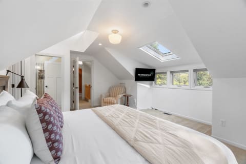 Bright and airy attic bedroom featuring a bed, chair, and television, with a skylight and windows.