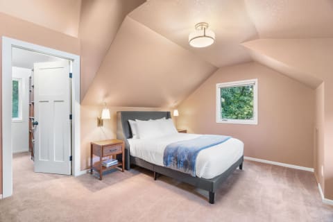 Interior view of a modern attic bedroom with a sloped ceiling, a bed with white linens, and a green view outside.