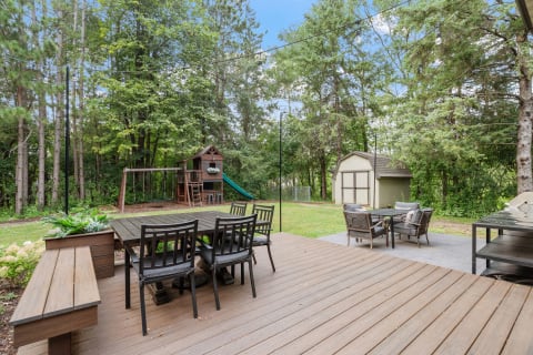 Outdoor wooden deck featuring a dining table, chairs, a play structure, and a shed in a green backyard.