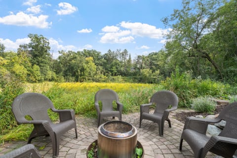 An outdoor fire pit with chairs on a stone patio, overlooking a field and trees under a blue sky.