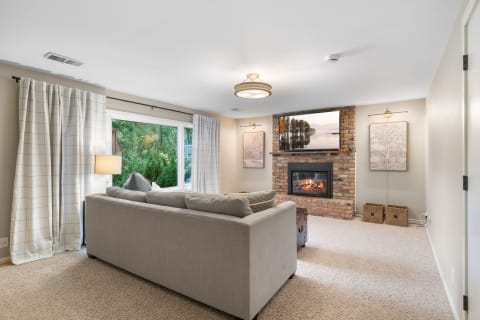 A cozy living room with a sectional sofa, brick fireplace, and large window with curtains.