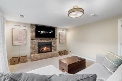 A contemporary living room with a gray sofa, a brick fireplace, and a mounted TV displaying 'MINNESTAY'.