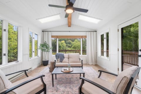 Sunroom with a sofa, chairs, and large windows showcasing green foliage outside.