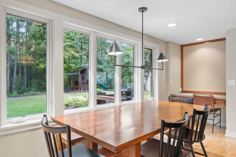 Interior view of a dining area with wooden table and chairs, large windows, and a view of the garden.