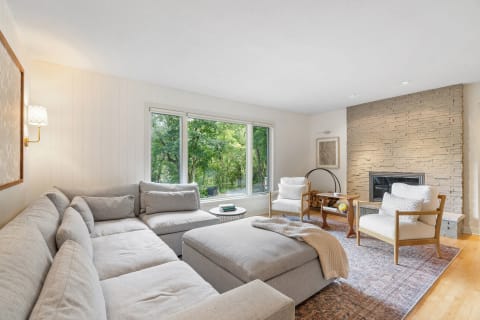 A living room featuring a gray sectional sofa, natural light from large windows, and a stone fireplace.