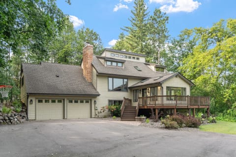 A charming house surrounded by trees, featuring a wooden deck, stone chimney, and basketball hoop.