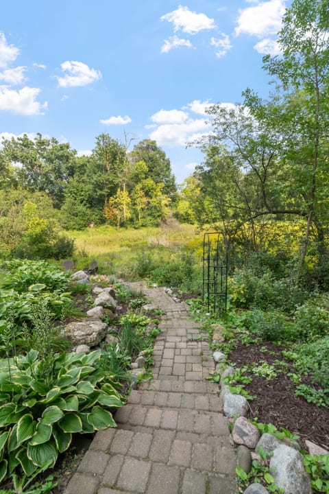 A cobblestone garden path surrounded by lush plants and trees under a clear sky.