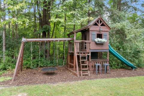 A wooden playset with a house, slide, and swings in a green backyard.