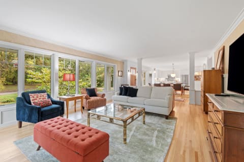 A spacious living room with a beige sofa, colorful ottoman, and large windows.