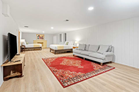 A spacious, well-lit basement room featuring a gray sofa, two beds, a red area rug, and a TV.