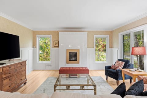 Modern living room featuring a fireplace, navy chair, and red ottoman with natural light streaming in through windows.