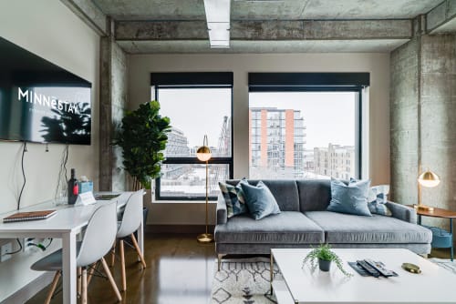 Unwind in your stylish industrial loft with exposed concrete beams, plush gray seating, and stunning city views through floor-to-ceiling windows.